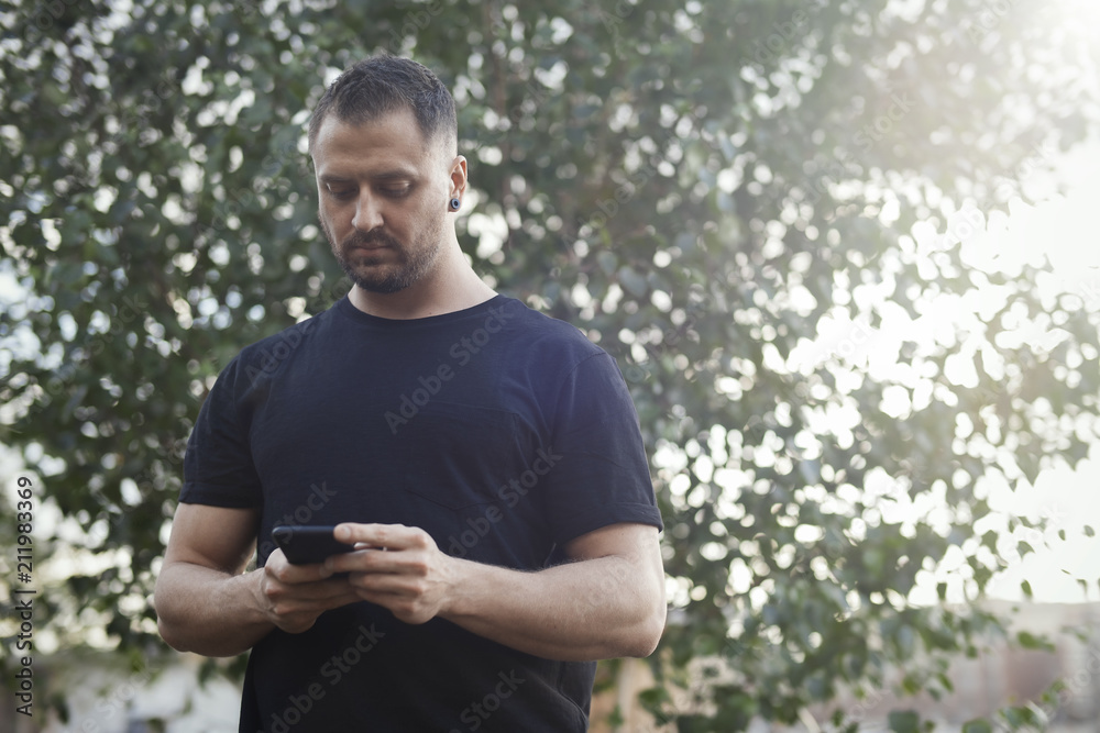 Fototapeta premium Young man typing text message on mobile phone outdoors in the park.