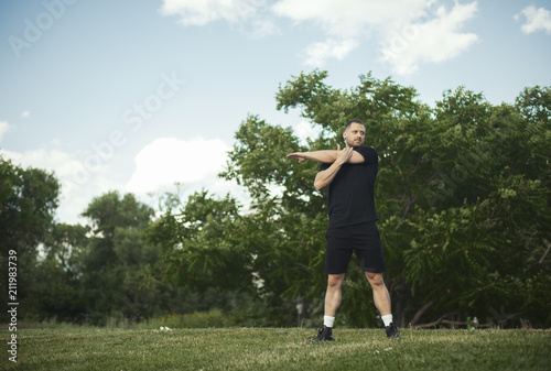 Distant plan of attractive man doing stretching hands exercises, in park.