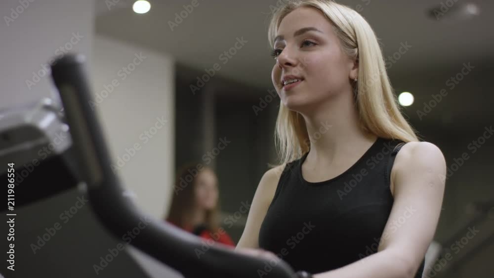 blonde girl turns the pedals on an exercise bikeand smiles at the other trainer in new gym blonde girl turns the pedals on an exercise bikeand smiles at the other trainer in new gym