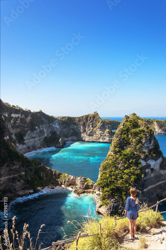 Young happy girl looking amazing view of a tropical beach, sea rocks and turquoise ocean, blue sky. Atuh beach, Nusa Penida island, Indonesia. Travel and adventure background. photo
