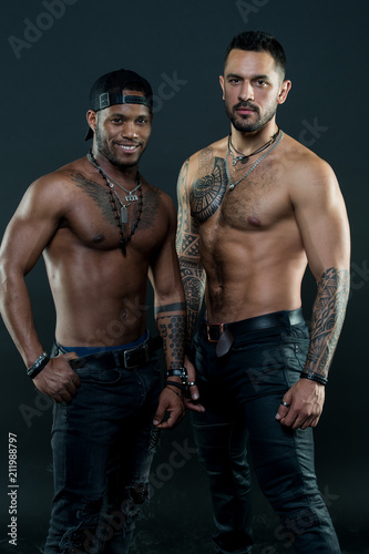 Friends with bare torsos wearing dark jeans. African and Caucasian men posing for fashion campaign. Bodybuilders with tattooed arms and chests on black background, sport and body care concept