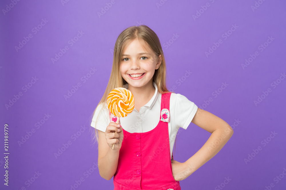 Smiling child with bright lollipop. Girl with long blond hair in pink outfit isolated on purple background. Sweet tooth with happy face holding big round candy, dessert time concept Smiling child with bright lollipop. Girl with long blond hair in pink outfit isolated on purple background. Sweet tooth with happy face holding big round candy, dessert time concept