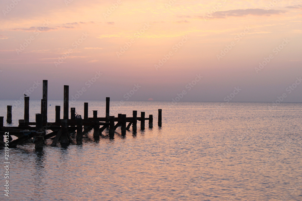 Sunrise on the sea. The sky is painted in shades of pink. Wooden piles of destroyed pier sticking out of the water.