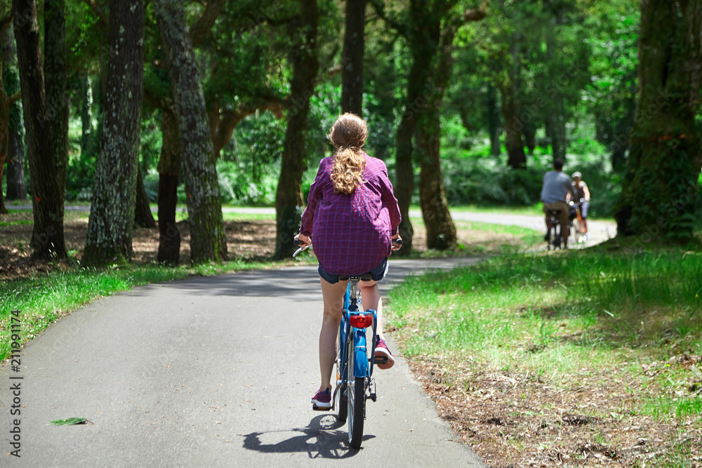 Young woman is riding a blue bicycle in a purple shirt in the park. View from the back. Bike ride in the nature on a sunny summer day Young woman is riding a blue bicycle in a purple shirt in the park. View from the back. Bike ride in the nature on a sunny summer day