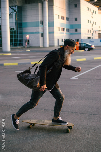 Stylish handsome middle-aged man with long gray beard riding on longboard during sunset on urban background