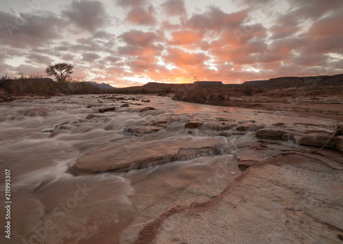The Virgin river flows over sandstone ledges while the morning sky reflects the orange clouds of sunrise.