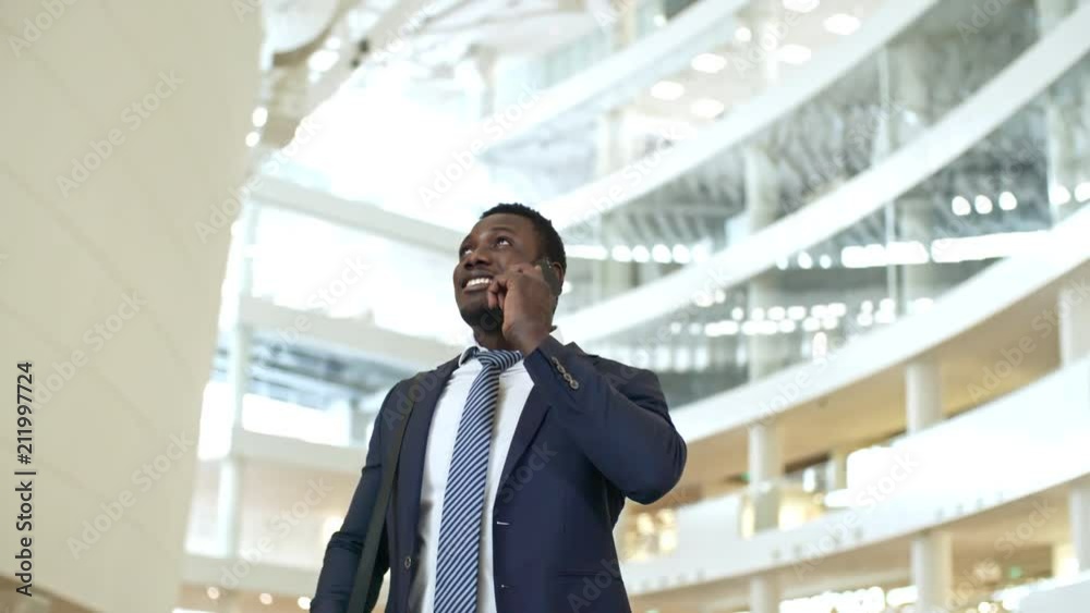 PAN shot of African American businessman in formal suit standing in modern office center, looking up and talking on cell phone PAN shot of African American businessman in formal suit standing in modern office center, looking up and talking on cell phone
