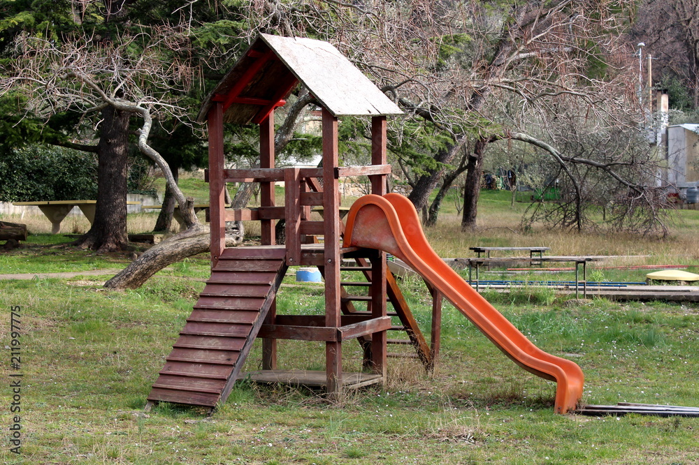 Outdoor wooden public playground equipment with climbing steps and plastic slide surrounded with winter vegetation and uncut grass