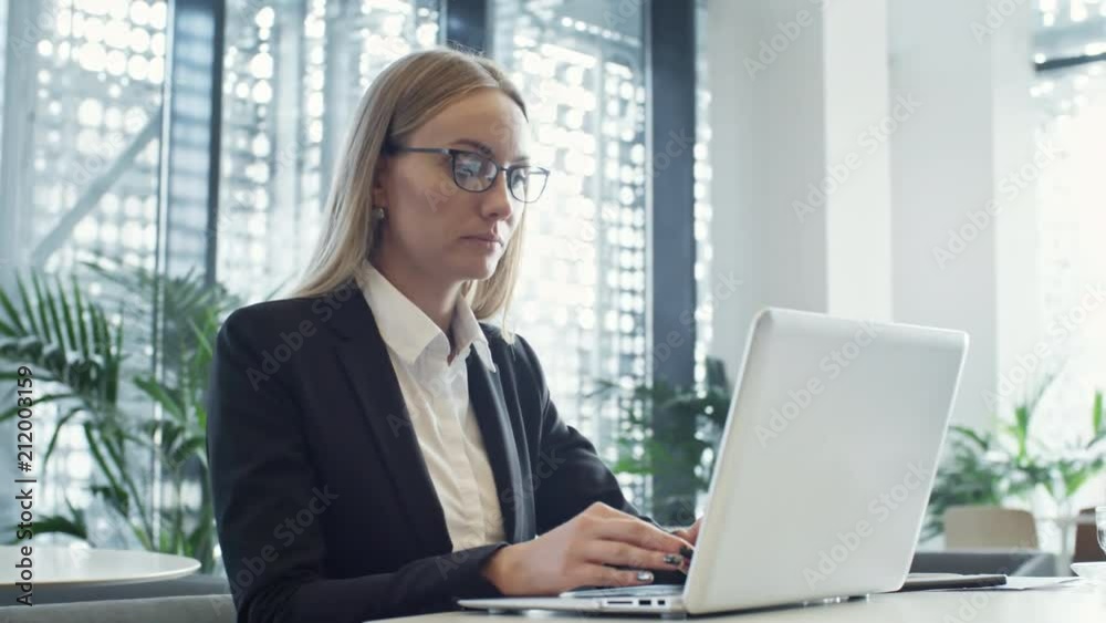 Panning shot of beautiful blonde businesswoman in eyeglasses typing on laptop in modern coworking space; two businessman taking selfie in background Panning shot of beautiful blonde businesswoman in eyeglasses typing on laptop in modern coworking space; two businessman taking selfie in background