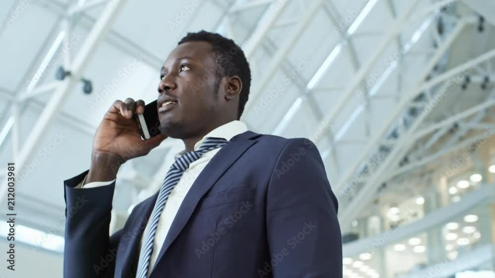 PAN low-angled shot of African American businessman in formal suit standing in office center and speaking on mobile phone with client PAN low-angled shot of African American businessman in formal suit standing in office center and speaking on mobile phone with client