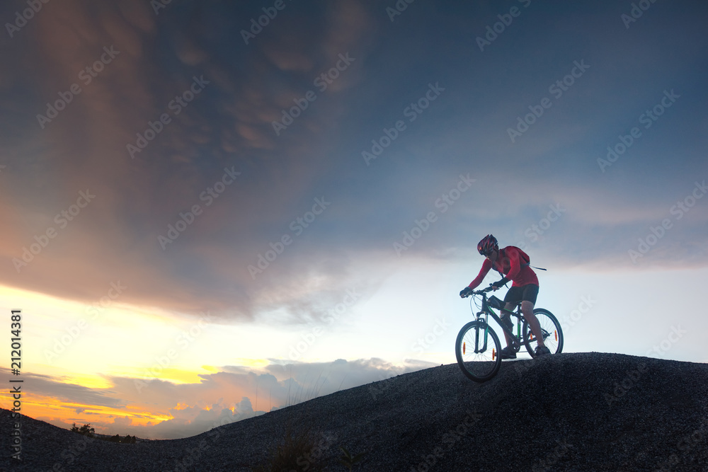 man cyclist biker riding mountain bike running from the hill at sunset in background