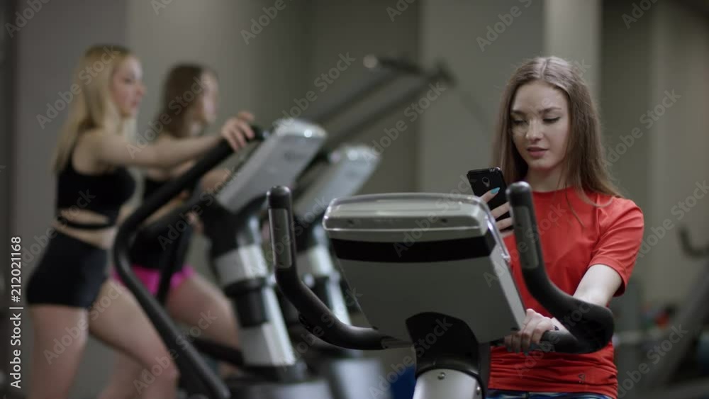 awesome girl in red shirt vigorously work on exercise bike and take phone to chek mail in the new gym against the background of engaged girls awesome girl in red shirt vigorously work on exercise bike and take phone to chek mail in the new gym against the background of engaged girls