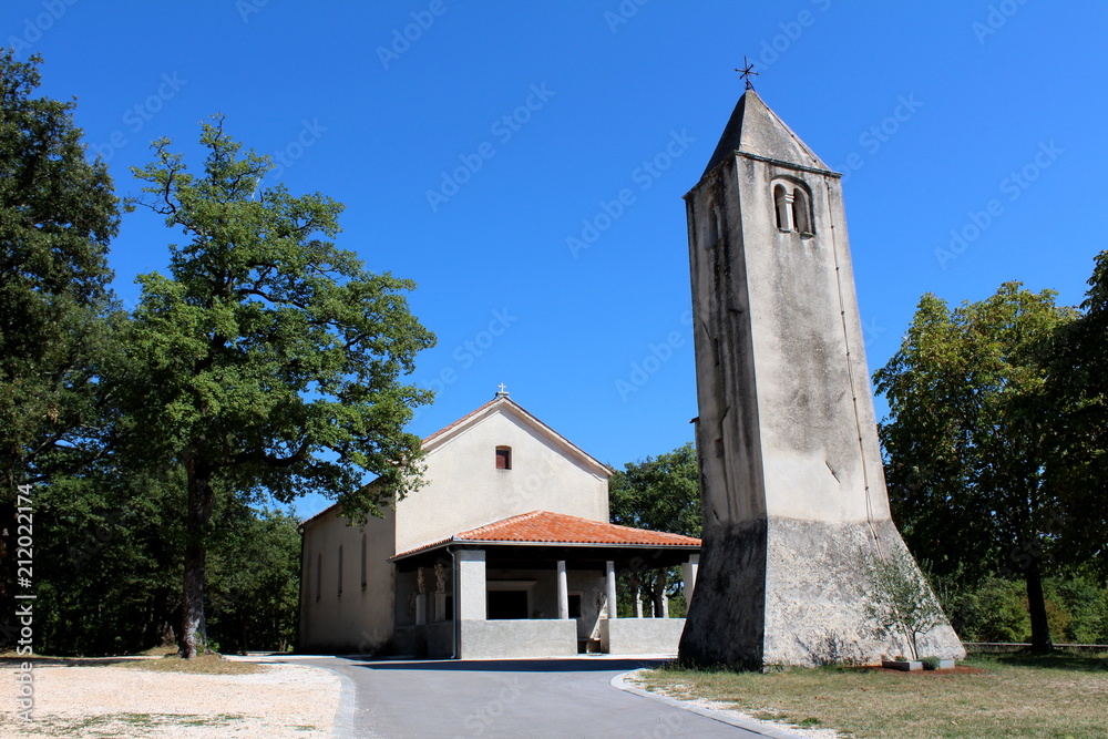 Stone bell tower with old renovated church in background surrounded with trees and paved road