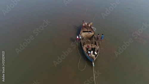 Aerial view. Flying around a small boat on which several Indian workers extract sand from the bottom of the muddy river by hand, with the help of sticks, buckets and ropes.  photo