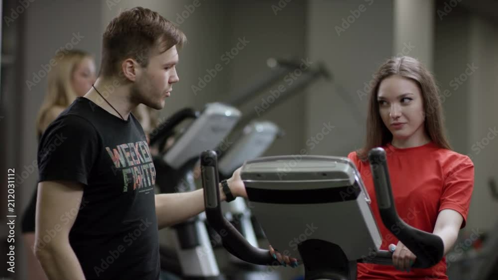 nice girl in red shirt vigorously work on exercise bike and coach come instructs on the correct execution of the exercise in the new gym against the background of engaged girls nice girl in red shirt vigorously work on exercise bike and coach come instructs on the correct execution of the exercise in the new gym against the background of engaged girls