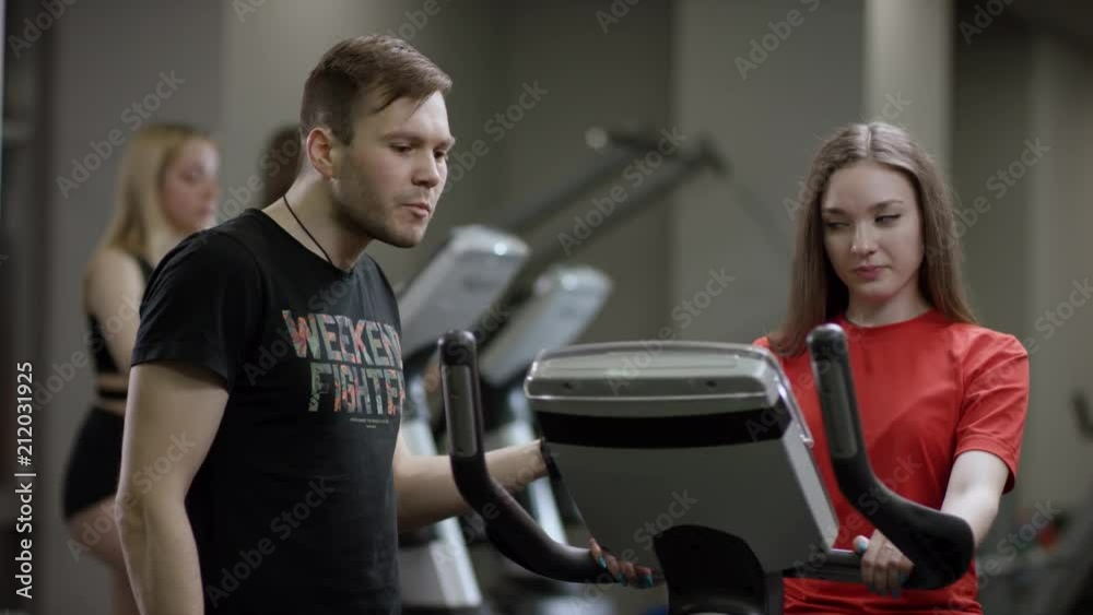 beautiful girl in red shirt vigorously work on exercise bike and coach come instructs on the correct execution of the exercise in the new gym against the background of engaged girls beautiful girl in red shirt vigorously work on exercise bike and coach come instructs on the correct execution of the exercise in the new gym against the background of engaged girls