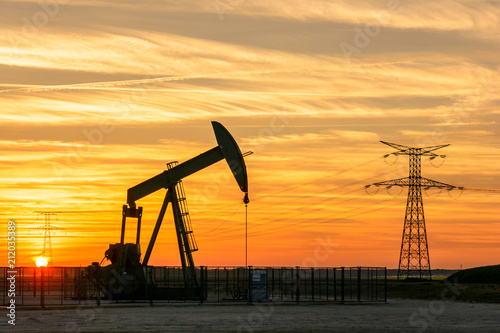 View of a pumpjack at sunset pumping oil out of a well with the silhouettes of transmission towers supporting an overhead power line in the background against a red sky. photo