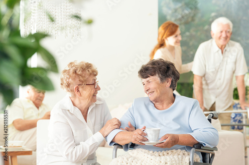 Front view of two happy geriatric women talking and holding hands in a private luxury day care center. Other seniors in the blurred background.