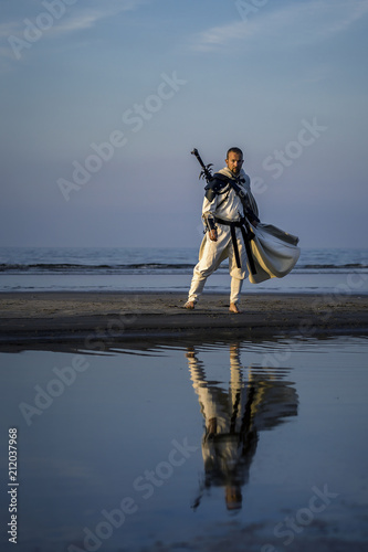Portrait of assassin in white costume with the sword at the sea. He is posing...