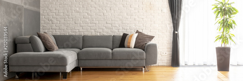 Grey corner sofa with decorative cushions in the real photo of bright living room interior with brick wall, fresh green plant and window with drapes