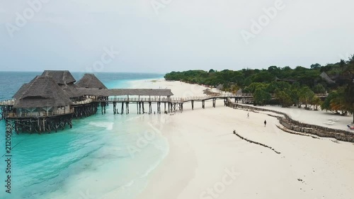 Flying towards wooden house with thatched roof and pier bridge on stilts. Wide paradise beach, white sand, azure ocean on cloudy day. Nungwi and Kendwa beach coastline, Zanzibar, Tanzania.