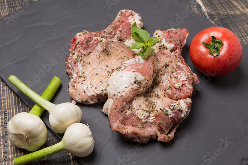 two pieces of steak with spices on black slate, three heads of garlic and tomato, raw meat and green basil leaves, top view