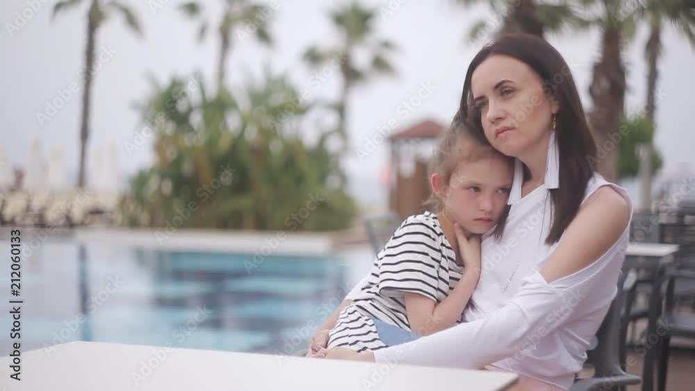 Mother and sad daughter sitting at a table by the pool Mother and sad daughter sitting at a table by the pool