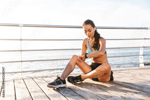 Photo of brunette athlete sportswoman 20s in sneakers looking at wrist watch, while sitting on wooden floor of boardwalk in sunny morning with sea background
