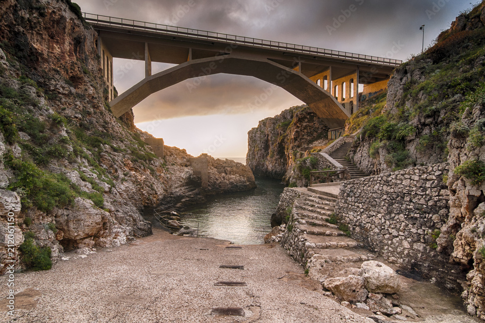 Italia, Luglio 2018 - alba al ponte Ciolo tra Santa Maria di Leuca e Otranto. Si nota il ponte illuminato dal sole e la piccola spiaggia deserta Italia, Luglio 2018 - alba al ponte Ciolo tra Santa Maria di Leuca e Otranto. Si nota il ponte illuminato dal sole e la piccola spiaggia deserta