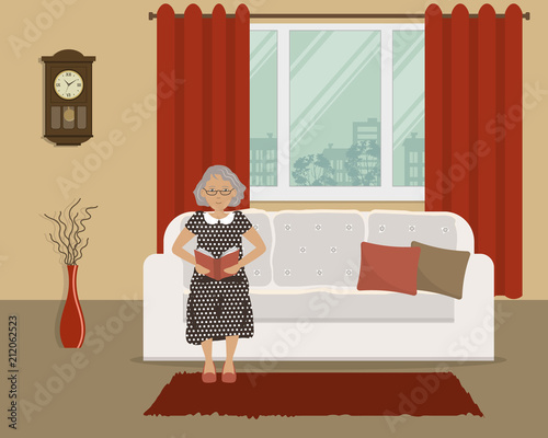 Elderly lady is sitting on the couch and reading a book. There is a white sofa on the window background in the living room. There is also a wall clock and a vase with decorative branches in the room