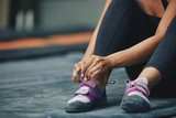 Crop sportive female putting on sneakers sitting on mat in gym on blur background