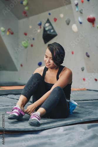 Asian brunette female in sportswear and insurance equipment on belt sitting on mat putting on trainers in climbing club
