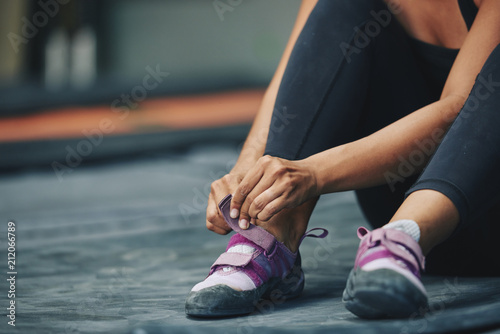 Crop sportive female putting on sneakers sitting on mat in gym on blur background