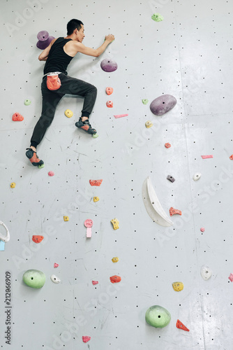 From below view of fit man climbing wall with colorful boulders in special modern gym