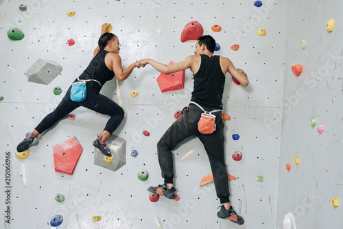 Back view man and woman working out in team and hitting hands while hanging on wall in bouldering center