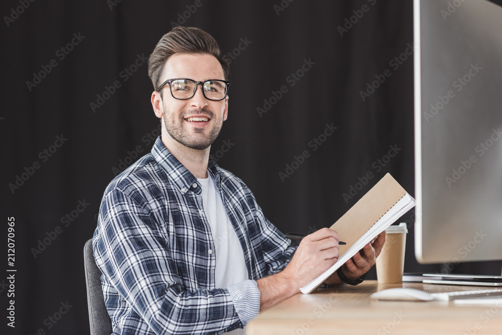 handsome young man in eyeglasses taking notes in notebook and smiling at camera while sitting at workplace handsome young man in eyeglasses taking notes in notebook and smiling at camera while sitting at workplace