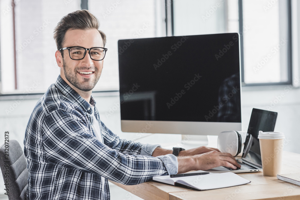handsome young programmer in eyeglasses smiling at camera while working with laptop and desktop computer handsome young programmer in eyeglasses smiling at camera while working with laptop and desktop computer