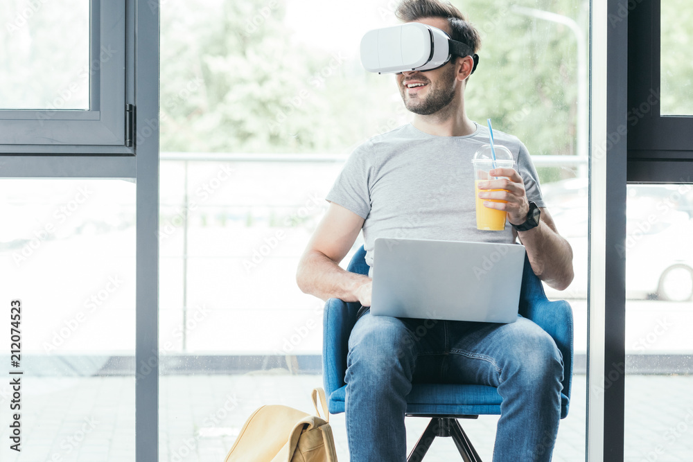 smiling young man holding plastic cup with fresh juice and using virtual reality headset with laptop smiling young man holding plastic cup with fresh juice and using virtual reality headset with laptop
