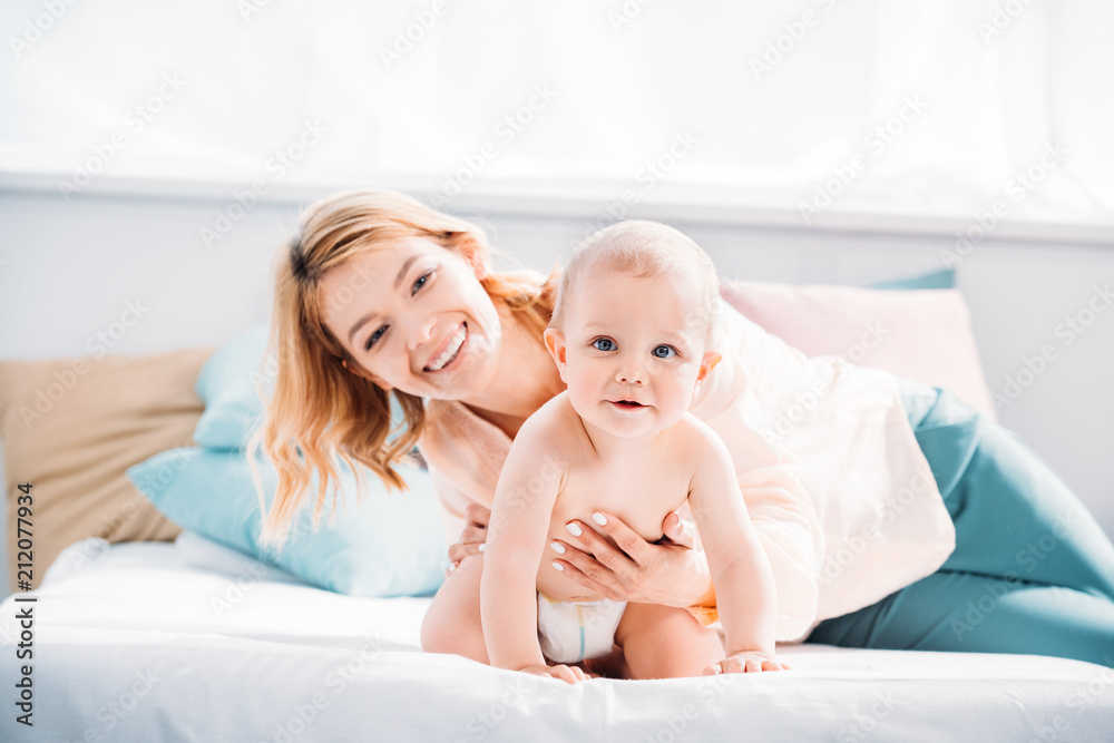 mother supporting her little child while he crawling on bed at home mother supporting her little child while he crawling on bed at home
