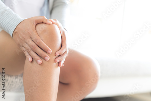 Closeup young woman sitting on sofa and feeling knee pain and she massage her knee at home. Healthcare and medical concept. photo