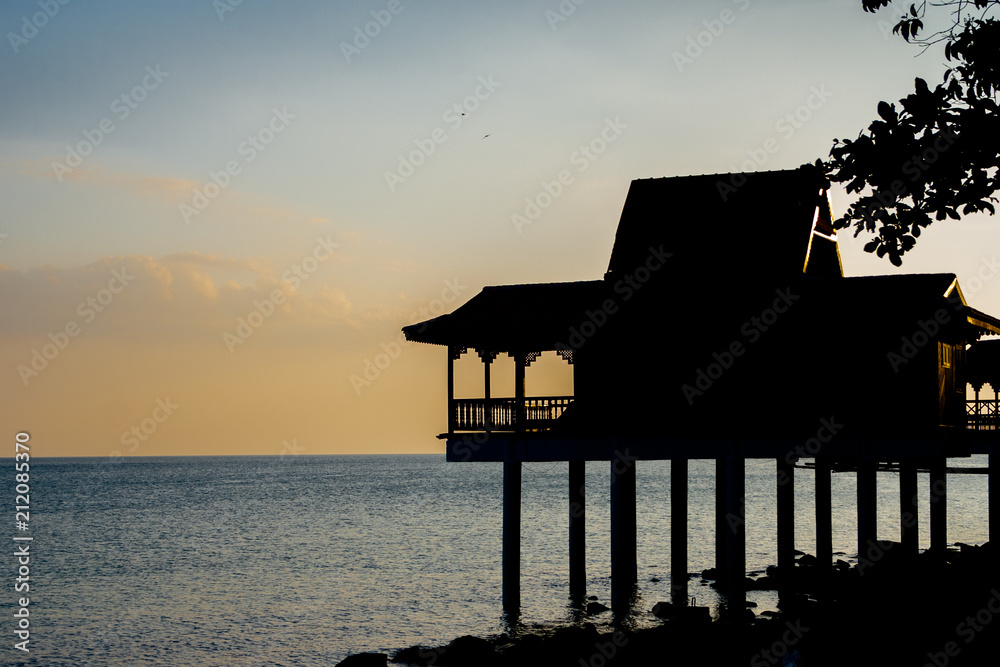 A picturesque scene of the black silhouette of a traditional Malay house on stilts with a tropically-suited roof at the sea during sunset in Malaysia.
