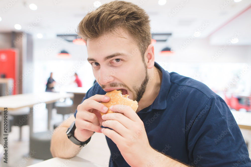 Handsome man sits in a fast food restaurant, bites a burger and looks at the camera. Lunch is a bad fast food. Fastfood concept. student eats a burger in a fast food restaurant. Handsome man sits in a fast food restaurant, bites a burger and looks at the camera. Lunch is a bad fast food. Fastfood concept. student eats a burger in a fast food restaurant.