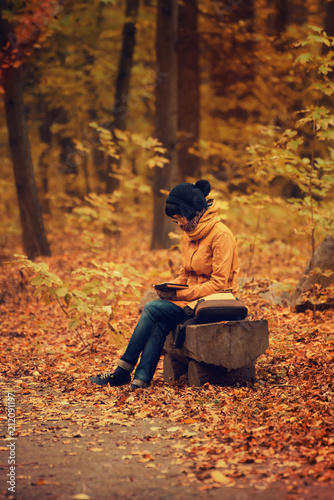 Young stylish hipster girl sitting on the bench in the autumn park and reading electronic book