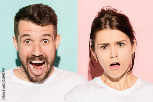 Closeup portrait of young couple, man, woman. One being excited happy smiling, other serious, concerned, unhappy on pink and blue background. Emotion contrasts