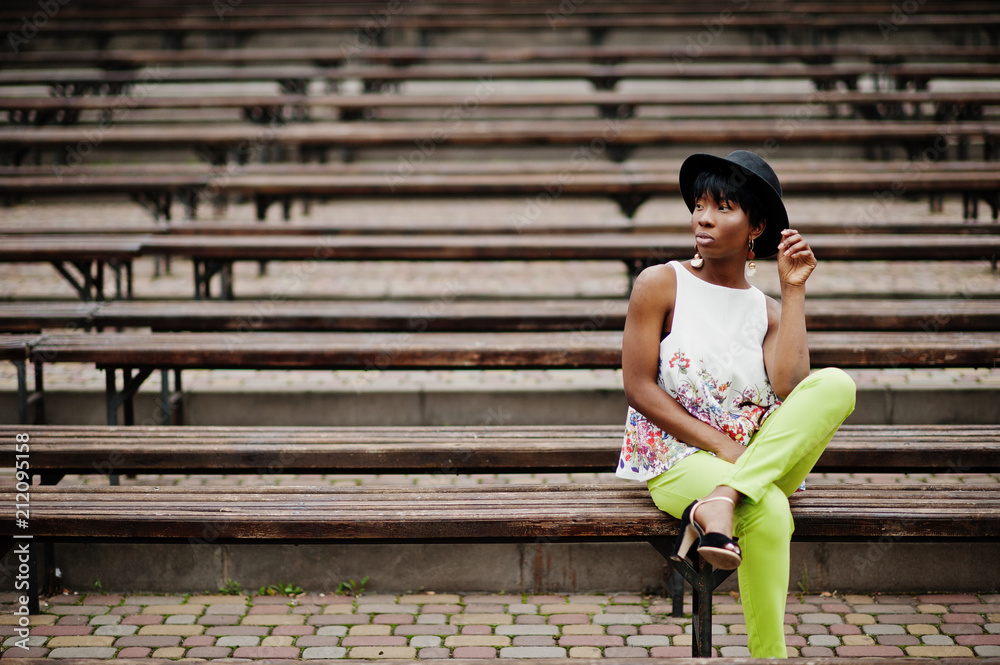 Amazing african american model woman in green pants and black hat posed at bench.