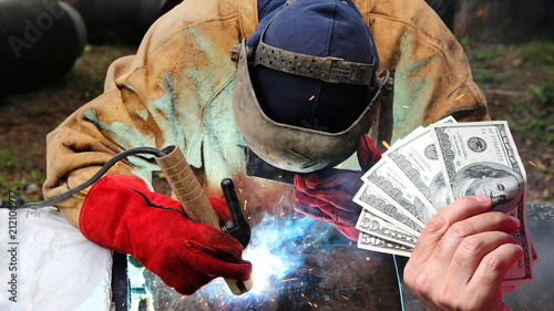 Pipeline Welder Salaries As A Concept / Hand holding US dollar bills in front of an pipeline welder at work with protective equipment