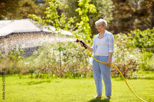 gardening and people concept - happy senior woman watering lawn by garden hose at summer