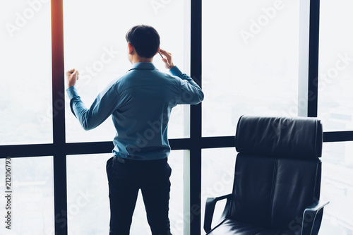 Happy businessman standing alongside large windows, talking on his mobile phone and looking out