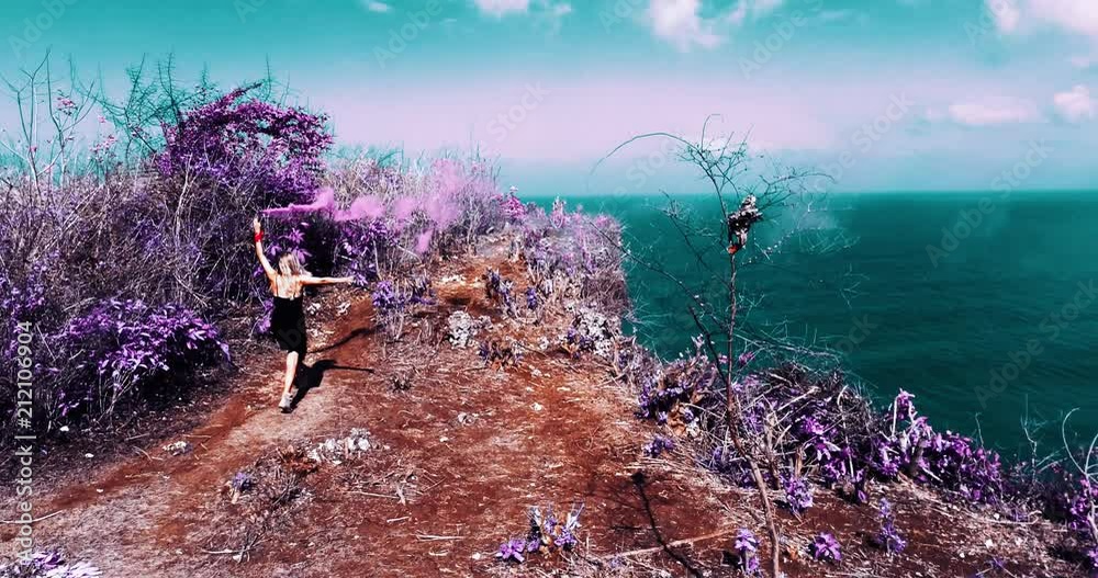 Aerial drone shot of pretty girl playing  with colored smoke on the cliff with amazing ocean and cloudy sky view - video in slow motion
