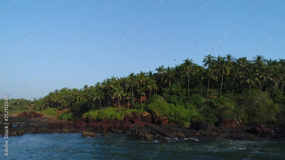 Aerial view. Thickets of coconut palms on the shore of an uninhabited tropical island. Waves breaking on coastal reefs.The camera slowly glides at a low altitude. Paradise landscape. Aerial view. Thickets of coconut palms on the shore of an uninhabited tropical island. Waves breaking on coastal reefs.The camera slowly glides at a low altitude. Paradise landscape.